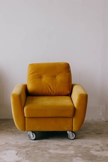 A mustard yellow upholstered armchair with a cushioned backrest and armrests, situated on a caster wheel base on a concrete floor against a plain white wall. The chair appears clean, with smooth fabric, and the area around is minimalistic without additional objects or decorations. This setting illustrates a simple domestic or commercial environment emphasizing surface cleanliness, which is relevant to proper upholstery surface cleaning and maintenance. For effective deep cleaning and sanitisation of similar furniture, Carpet Cleaning Harringay offers professional services to avoid common upholstery cleaning mistakes made by landlords. The overall scene highlights the importance of regular cleaning to maintain hygiene and prolong furniture life.