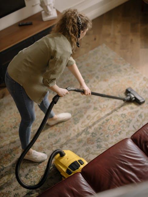 A woman vacuuming a patterned area rug in a living room, using a yellow canister vacuum cleaner. She is dressed casually in a light green jacket, dark jeans, and white sneakers, and is leaning forward slightly to clean thoroughly. The room features wooden flooring, a leather sofa in the foreground, and a wooden sideboard with decorative items along the wall. Natural lighting illuminates the scene, highlighting the cleanliness and maintenance of the carpet, consistent with professional deep cleaning services by Carpet Cleaning Harringay. The setting emphasizes surface cleaning and sanitisation of domestic living spaces.