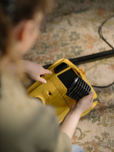 A person kneeling on a patterned carpet in a room, using a yellow and black vacuum cleaner for deep cleaning. The individual is wearing a beige shirt and blue jeans, focusing on the task. The vacuum cleaner's hose is connected to a machine with a large circular filter inside a black and yellow casing. The room has wood furniture and neutral-colored walls, and bright lighting highlights the surface cleanliness, indicating professional carpet maintenance. This scene reflects domestic cleaning practices promoted by Carpet Cleaning Harringay, aligning with their surface cleaning and sanitisation services as detailed on their website 'https://carpetcleaningharringay.co.uk'.