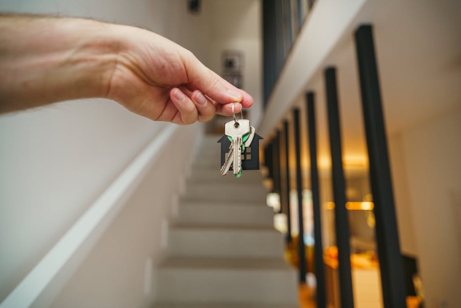A close-up of a human hand holding a set of keys, including a house key and a mailbox key, with a staircase and black railing visible in the background inside a modern residential building. The scene is well-lit with warm interior lighting, and the surfaces appear clean and well-maintained, consistent with professional domestic cleaning and surface sanitisation that Carpet Cleaning Harringay offers, in relation to the Haringey Council cleaning permits for Harringay landlords.