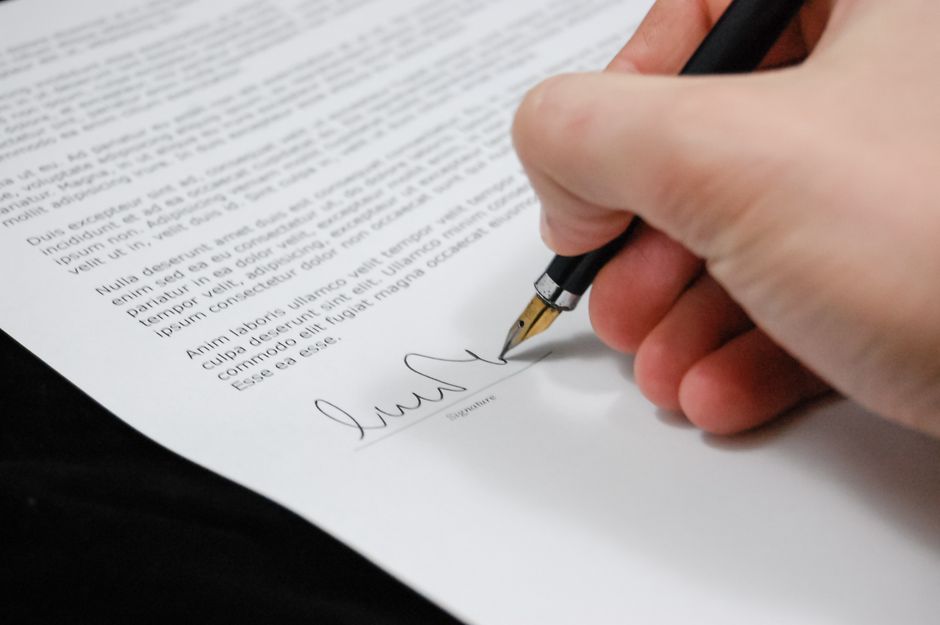 Close-up of a person's hand holding a black fountain pen and signing a document on a white sheet of paper, with printed text and placeholder Latin writing. The hand is positioned to the right, with visible fingers and thumb gripping the pen, which is poised over a signature line near the bottom of the page. The document rests on a smooth black surface, illuminated by soft, natural lighting, suggesting a professional environment. The setting reflects a focus on formalisation and cleanliness, aligned with professional surface cleaning and sanitisation practices carried out by Carpet Cleaning Harringay, relevant to the context of the Haringey Council cleaning permits for Harringay landlords.