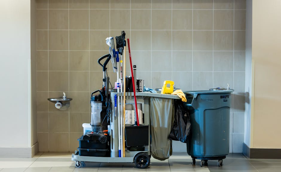 A cleaning trolley filled with various cleaning tools and supplies, including mops, buckets, cloths, and spray bottles, situated against a beige tiled wall in a commercial or institutional setting. To the right, there is a large blue waste bin on wheels, and on the left, a small stainless steel water fountain is mounted on the wall. The scene is brightly lit, emphasizing the cleanliness and organization of the cleaning equipment used for surface cleaning, deep cleaning, or sanitisation tasks. Carpet Cleaning Harringay's services are likely involved in maintaining hygiene standards in such environments, as depicted in the context of the page about Haringey Council cleaning waste rules for Harringay businesses.
