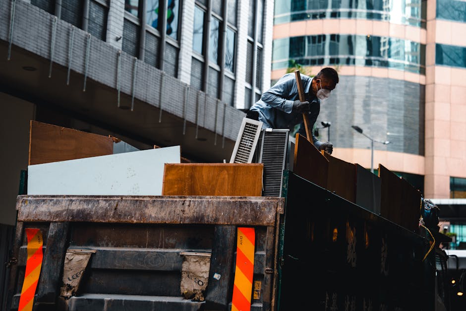 A man wearing a white face mask and protective gloves is standing inside the back of a large, open-top truck filled with various wooden furniture pieces, including cabinets and panels. The truck is parked outdoors in an urban setting with modern glass and brick office buildings in the background, under natural daylight. The man appears to be inspecting or arranging the furniture, which are likely destined for disposal or recycling, with a focus on proper waste management compliant with Haringey Council's cleaning waste rules for Harringay businesses. Visible tools include a broom or dustpan, and the furniture surfaces show signs of wear. The scene emphasizes surface cleaning and waste removal, reflecting professional commercial or domestic cleaning and sanitisation processes.