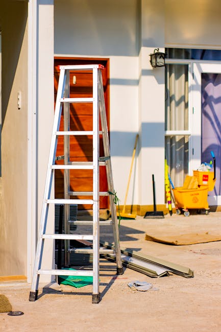 An indoor construction or renovation scene featuring a three-step aluminium ladder positioned on a smooth, light-colored floor. Behind the ladder, there is a partially finished wall with visible studs and a wooden door frame, indicating ongoing work. To the right, a window with open blinds allows natural light to illuminate the space, and a yellow mop bucket with cleaning supplies, including a mop and a squeegee, are placed nearby. The area shows signs of ongoing domestic cleaning or maintenance, with a few scattered tools and materials on the floor. Carpet Cleaning Harringay advises that regular surface cleaning and sanitisation of such areas enhance hygiene during home improvement projects.