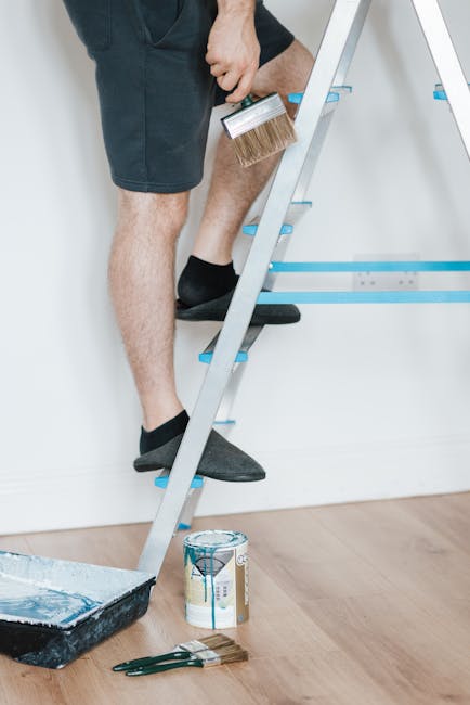 Close-up of a person standing on a silver and blue step ladder during domestic cleaning, holding a paintbrush in one hand with the bristles facing downward. The individual is wearing black shorts and black slip-on shoes. The scene is set in a well-lit interior with a white wall and wooden flooring. Below the ladder on the floor, there is a paint can with blue paint and a paint tray with some paint residue. The setting suggests preparation for surface cleaning or renovation. This image, associated with Carpet Cleaning Harringay, emphasizes the importance of thorough surface maintenance and deep cleaning for an end of tenancy or general hygiene effort.