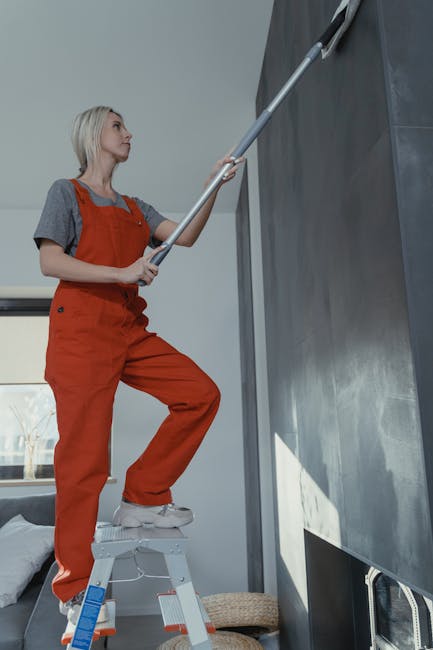 An indoor construction or renovation scene featuring a three-step aluminium ladder positioned on a smooth, light-colored floor. Behind the ladder, there is a partially finished wall with visible studs and a wooden door frame, indicating ongoing work. To the right, a window with open blinds allows natural light to illuminate the space, and a yellow mop bucket with cleaning supplies, including a mop and a squeegee, are placed nearby. The area shows signs of ongoing domestic cleaning or maintenance, with a few scattered tools and materials on the floor. Carpet Cleaning Harringay advises that regular surface cleaning and sanitisation of such areas enhance hygiene during home improvement projects.