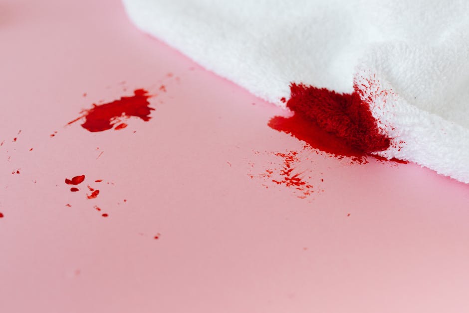 Close-up image of a white cloth wiping away fresh red wine spill stains on a smooth pink surface, likely a table or countertop. Visible red wine stains are scattered, with some streaks and droplets, indicating a recent spill. The white cloth is partially folded and positioned over part of the stain, suggesting an active cleaning process aimed at stain removal. The surface appears clean apart from the spill, and the lighting highlights the contrast between the red wine stains and the pink surface. This image relates to surface cleaning and stain removal services, as provided by Carpet Cleaning Harringay, emphasizing quick and effective domestic cleaning solutions.