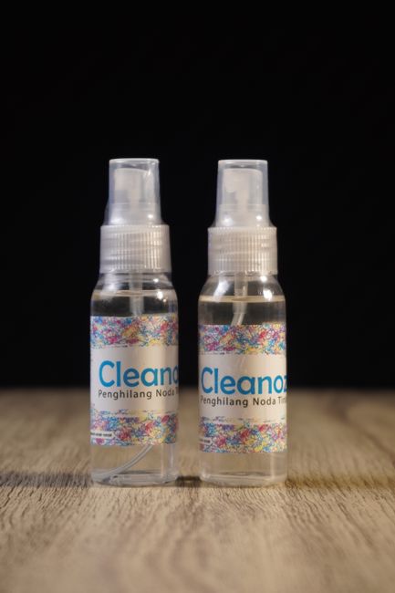 Two small transparent spray bottles filled with clear liquid are positioned side by side on a light wooden surface, against a black background. The bottles feature colorful, speckled labels with the text 'Cleanor' visible, indicating they are cleaning products. One bottle is slightly in front of the other, with both caps securely in place. The scene suggests a focus on surface cleaning solutions used for domestic or commercial cleaning tasks, aligning with the services offered by Carpet Cleaning Harringay for advanced and sanitising upkeep. The lighting highlights the bottles' transparency and the vibrant label design, emphasizing cleanliness and hygiene in a professional cleaning context.