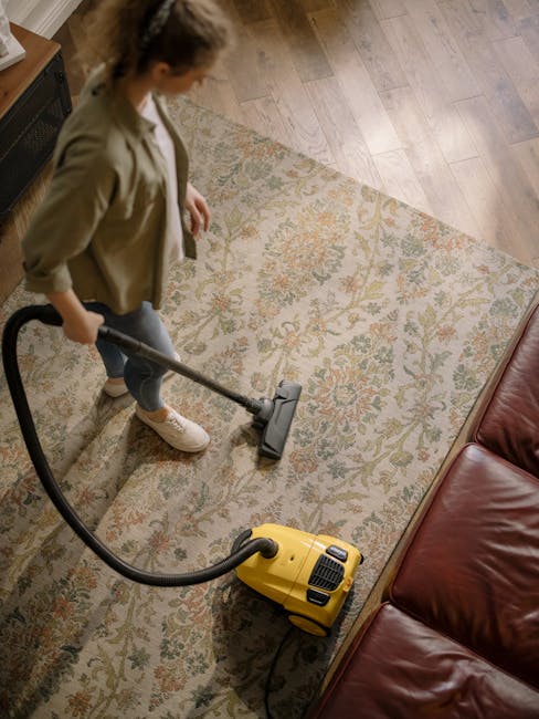 A woman with curly hair wearing a beige jacket, white top, and blue jeans using a yellow vacuum cleaner with a black hose to deep clean a patterned carpet in a living room. The carpet features a floral motif in soft tones and covers a wooden floor. Shelving and a leather sofa are visible, and the room is well-lit, highlighting the cleanliness achieved through professional surface cleaning by Carpet Cleaning Harringay. This image exemplifies domestic cleaning and hygienic maintenance.