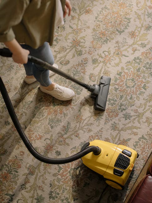 A woman with curly hair wearing a beige jacket, white top, and blue jeans using a yellow vacuum cleaner with a black hose to deep clean a patterned carpet in a living room. The carpet features a floral motif in soft tones and covers a wooden floor. Shelving and a leather sofa are visible, and the room is well-lit, highlighting the cleanliness achieved through professional surface cleaning by Carpet Cleaning Harringay. This image exemplifies domestic cleaning and hygienic maintenance.