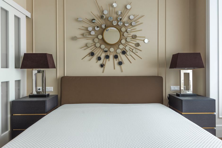 A neatly made bedroom with a beige upholstered headboard against a cream-colored wall featuring decorative vertical paneling. On either side of the bed, there are dark bedside tables topped with matching rectangular table lamps with dark brown shades and clear glass bases. Above the headboard hangs a round, sunburst-style mirror with a central circular mirror surrounded by radiating rods adorned with small circular accents of varying shades. The room is illuminated by natural light coming from a large window on the right, highlighting the clean, tidy surface of the white, textured bedspread. The overall scene exemplifies a hygienic and well-maintained space, suitable for professional domestic cleaning services by Carpet Cleaning Harringay.
