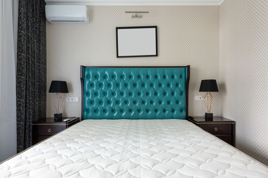 A neatly made hotel-style bedroom featuring a white mattress with a quilted surface, placed on a bed frame with an upholstered teal headboard. On either side of the bed are black bedside tables, each topped with a black table lamp with gold accents. The room has beige wallpaper with a subtle diamond pattern, and visible electrical outlets and switches. Mounted above the bed is a rectangular, black-framed mirror or artwork, and an air conditioning unit is installed high on the wall. The lighting is soft and even, enhancing the clean and hygienic appearance of the space, reflecting professional domestic cleaning standards maintained by Carpet Cleaning Harringay.