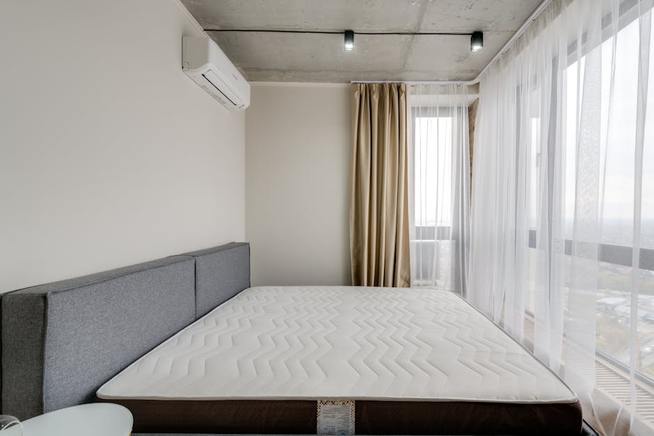 A neatly made bedroom with a beige upholstered headboard against a cream-colored wall featuring decorative vertical paneling. On either side of the bed, there are dark bedside tables topped with matching rectangular table lamps with dark brown shades and clear glass bases. Above the headboard hangs a round, sunburst-style mirror with a central circular mirror surrounded by radiating rods adorned with small circular accents of varying shades. The room is illuminated by natural light coming from a large window on the right, highlighting the clean, tidy surface of the white, textured bedspread. The overall scene exemplifies a hygienic and well-maintained space, suitable for professional domestic cleaning services by Carpet Cleaning Harringay.
